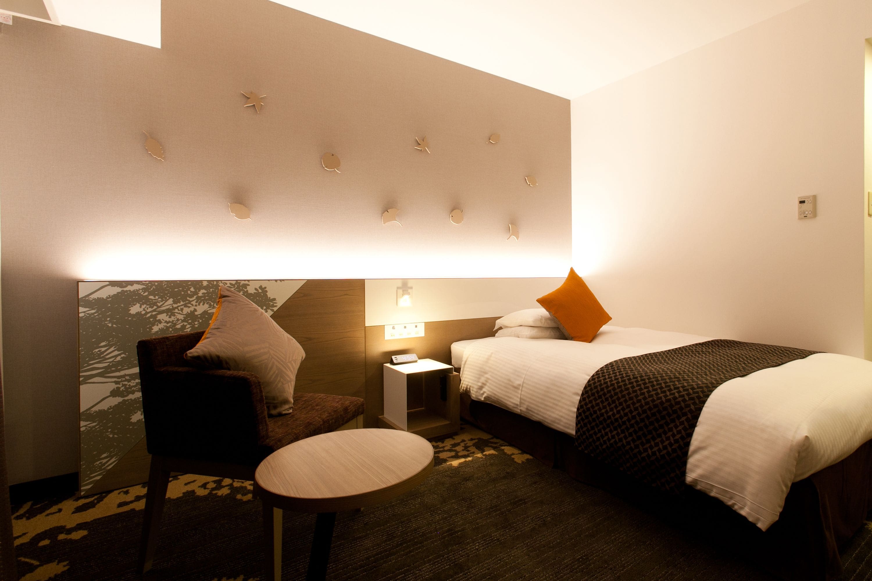 A hotel room with a bed, a chair, a small table, and a wall with decorative elements.