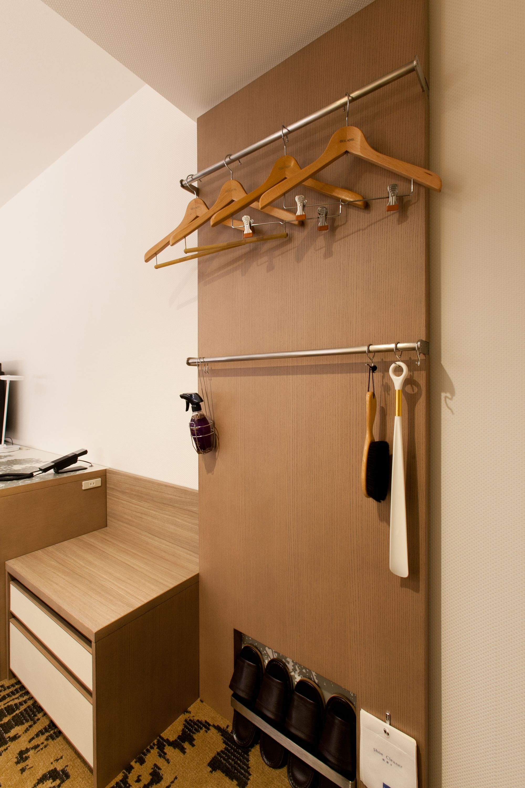A hotel room with a wooden bed, a built-in wardrobe with hanging space, a shoe rack, and a small table with a phone and remote control.