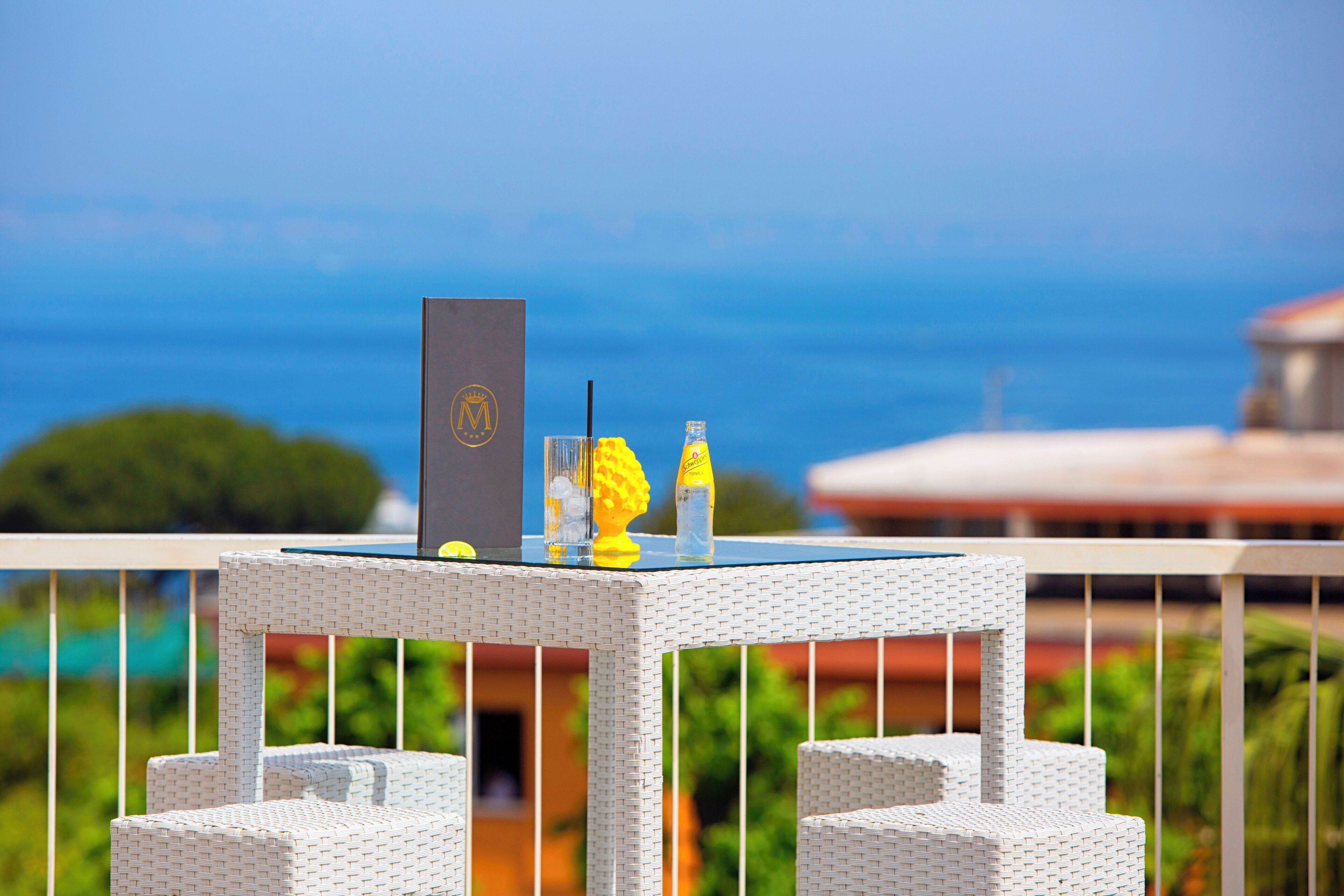 A wicker table with a bottle, a glass, and a box on a balcony overlooking a scenic view.