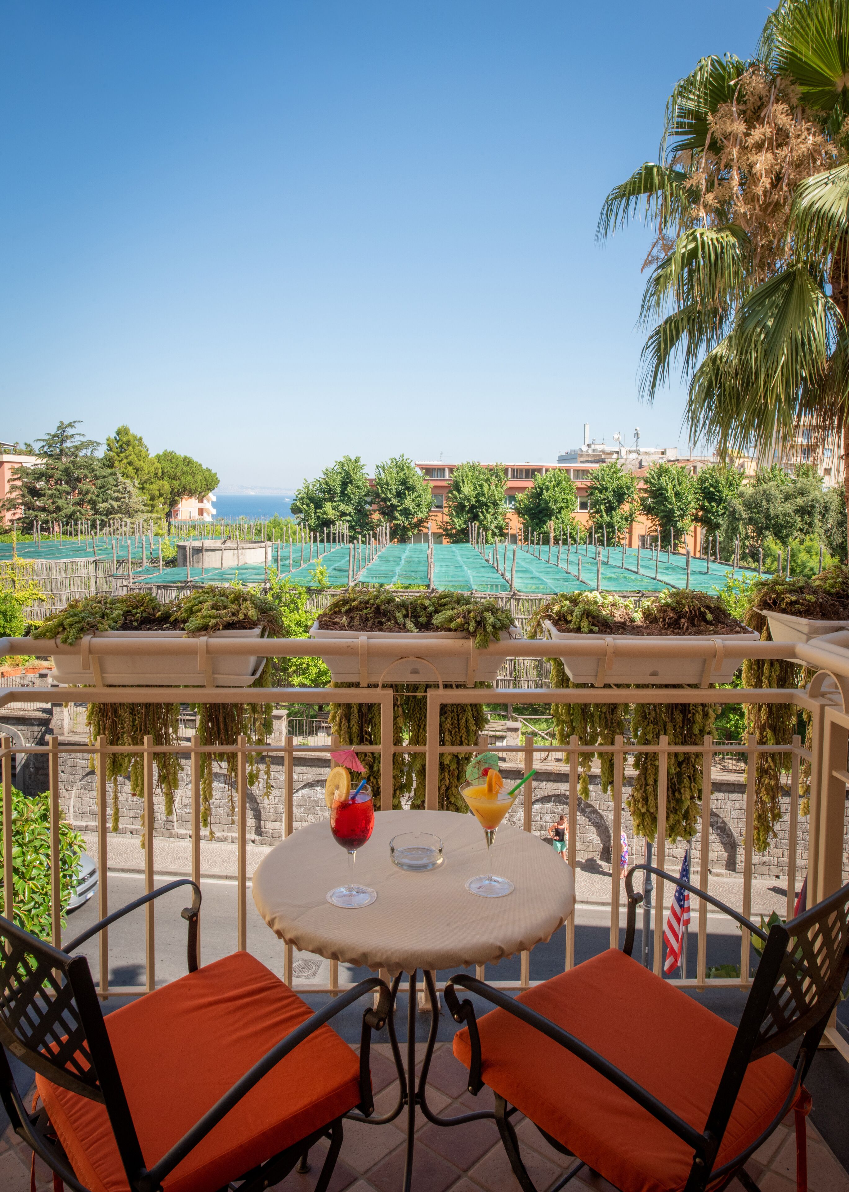 A balcony with two orange chairs and a table, overlooking a pool area with palm trees and buildings in the background.