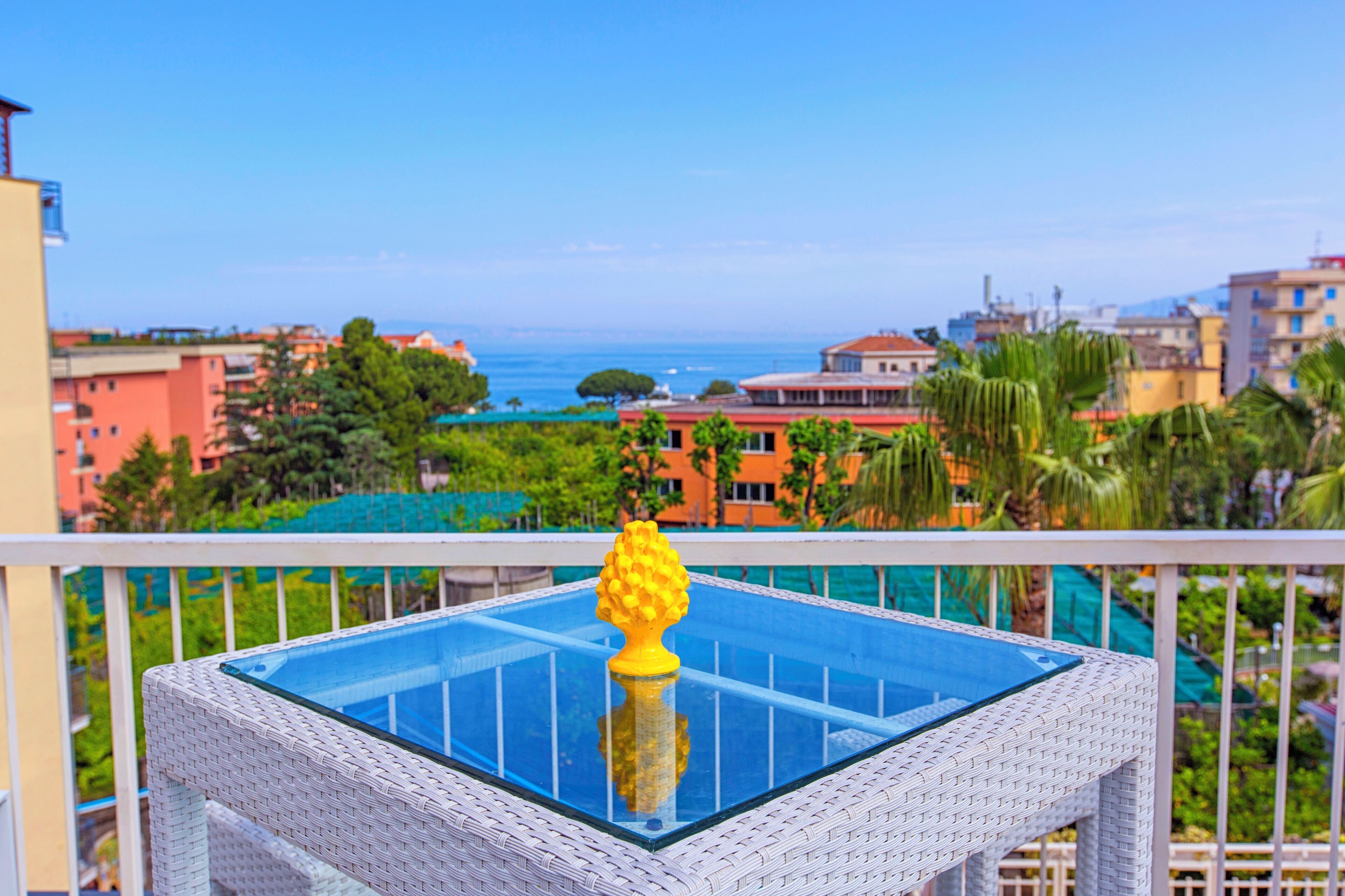 A balcony with a glass-top table, a yellow decorative object, and a view of the sea and buildings.