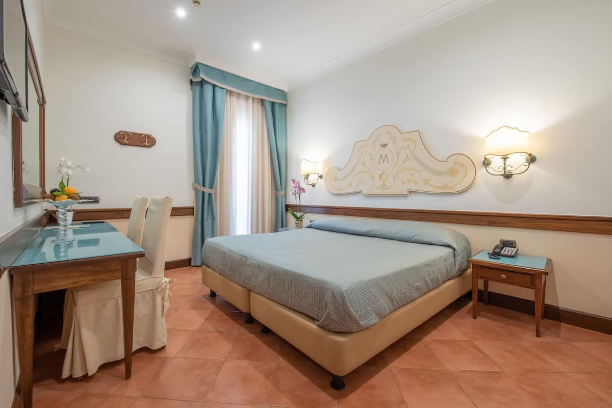 Une chambre d’hôtel comprenant un lit, un bureau, une chaise, une petite table, une table de chevet, un miroir et un luminaire fixé au mur.