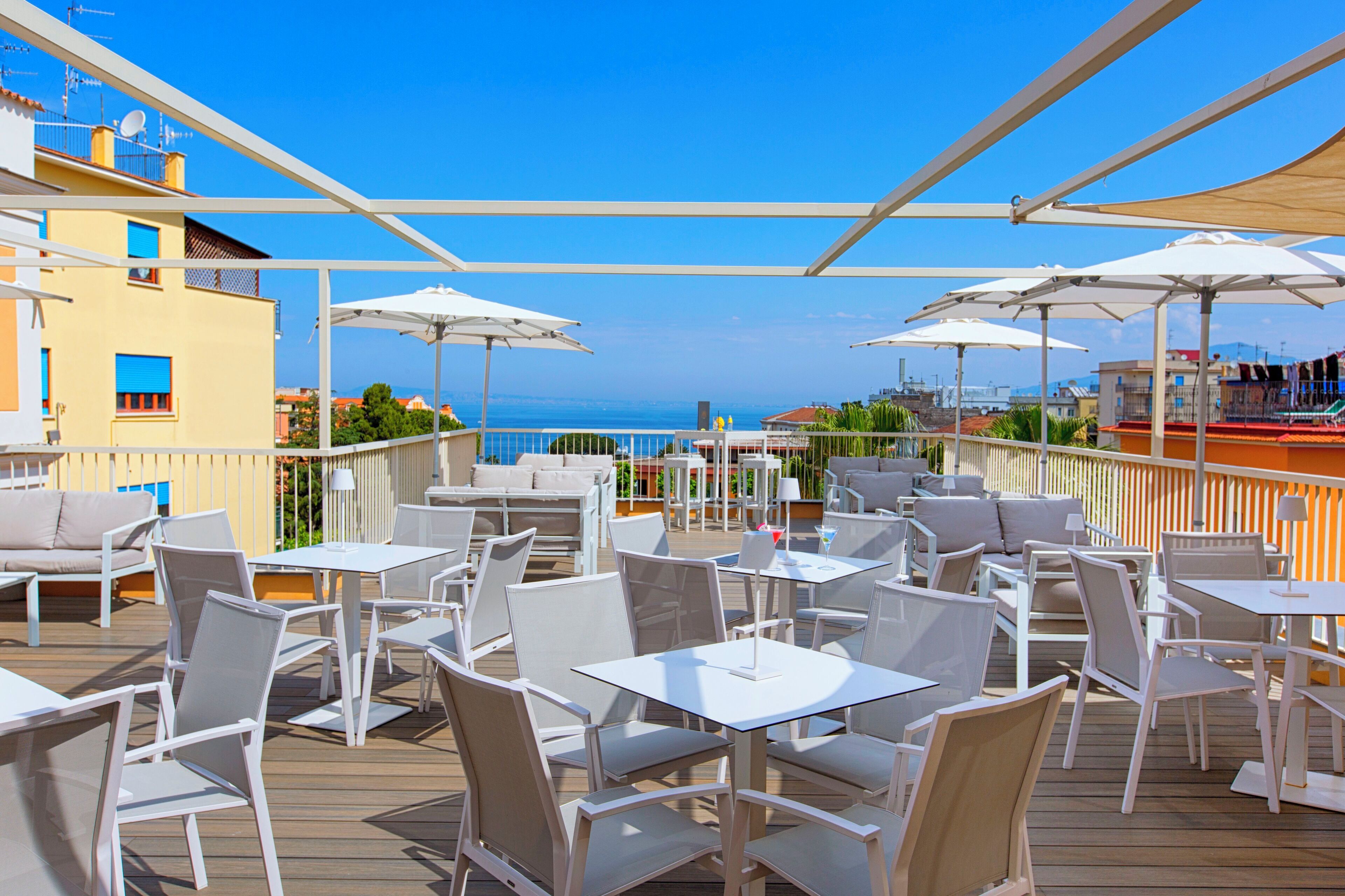 Una terrazza con tavoli e sedie bianche, ombrelloni e vista sul mare.