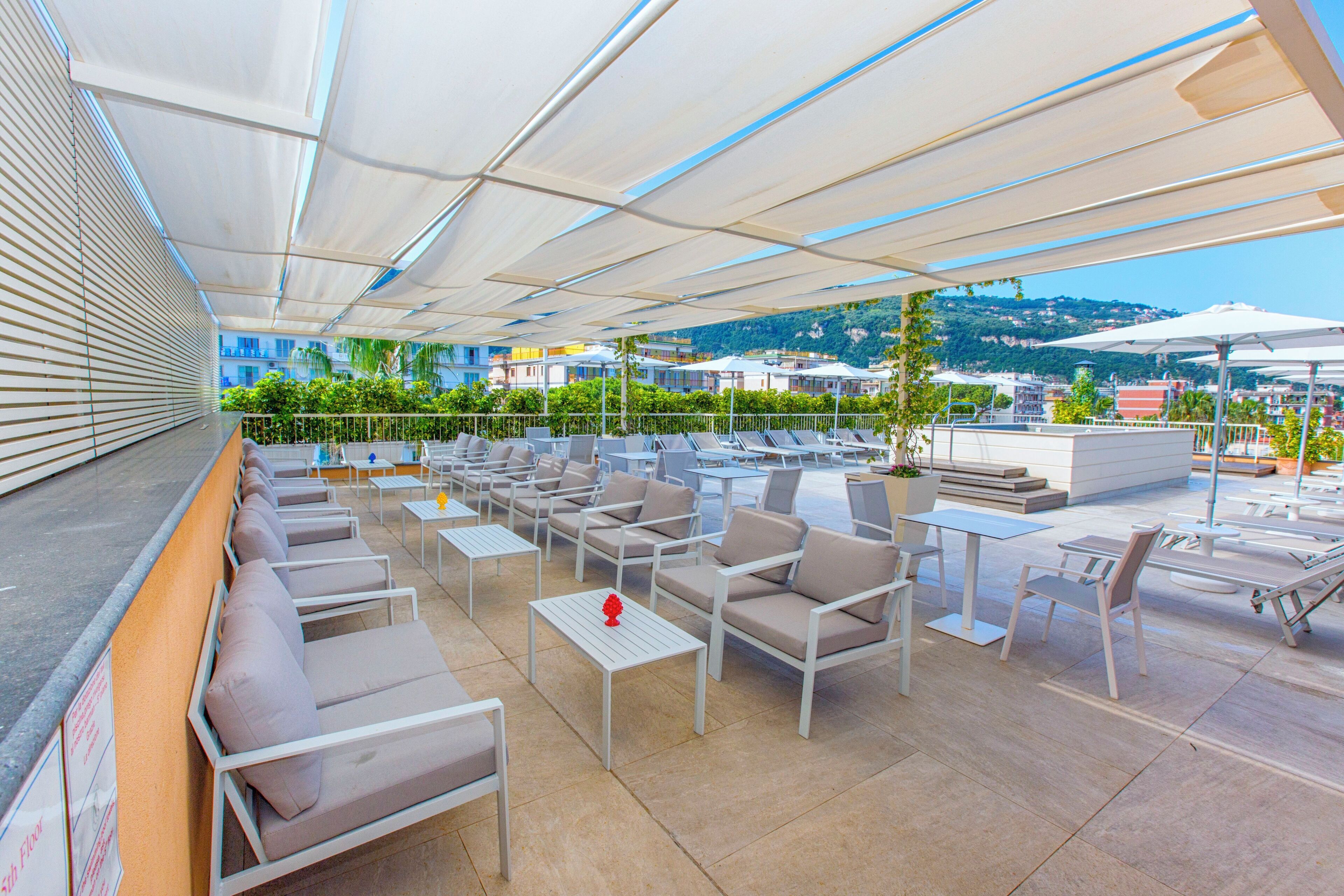 An outdoor seating area with multiple tables and chairs, a view of the city and hills in the background, and a covered section with umbrellas.