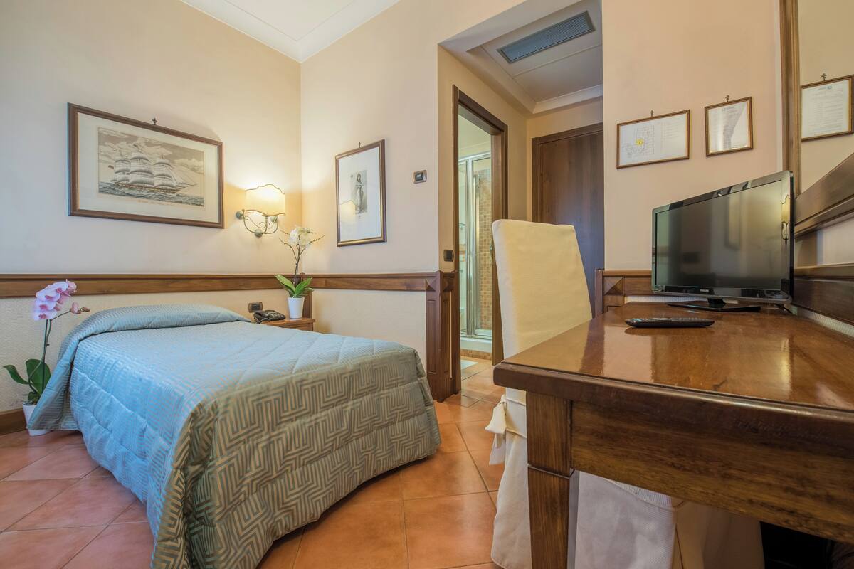 Une chambre d’hôtel avec un lit, un bureau avec une télévision, une chaise, une plante en pot et des tableaux encadrés au mur.