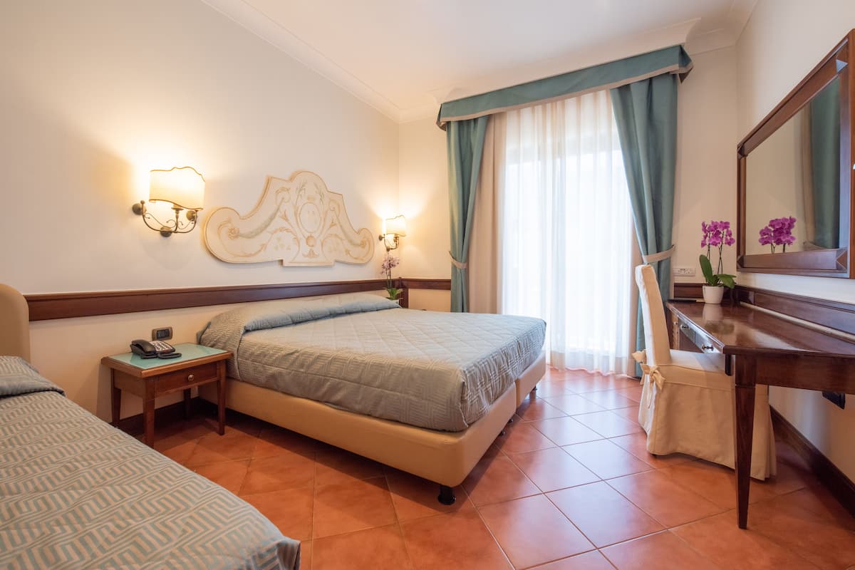 Une chambre d’hôtel comprenant un lit, une table de chevet avec un téléphone, un bureau avec un miroir et une fenêtre avec des rideaux.