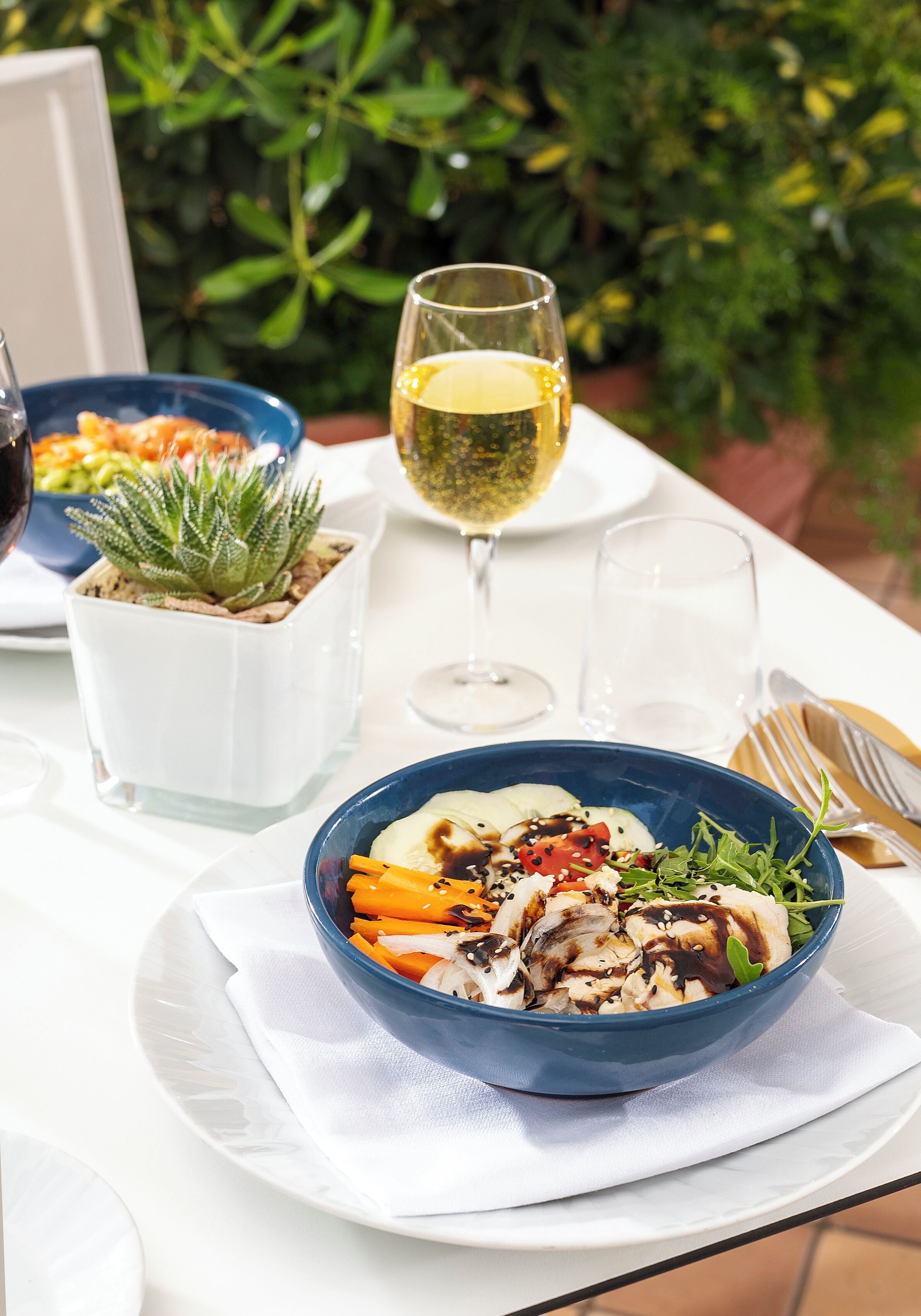 Ein Tisch gedeckt mit einer blauen Salatschüssel, einem Glas Weißwein und einem kleinen Topfpflanzen.