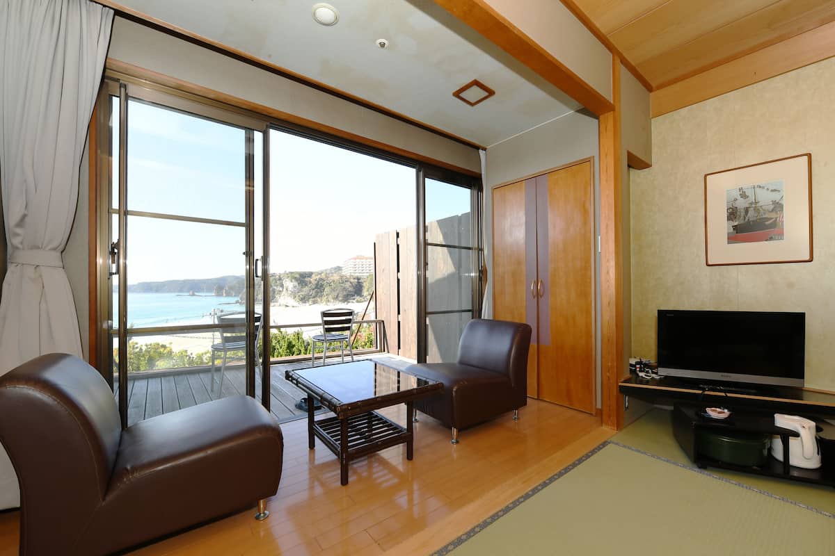 A room with a sliding glass door leading to a balcony with a view of the sea. There is a sofa, a small table, and a TV on a stand.
