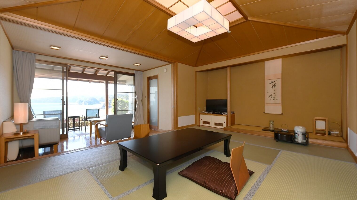 A traditional Japanese-style room with tatami flooring, a low table, a sofa, a television, and a sliding door leading to a balcony with a view.