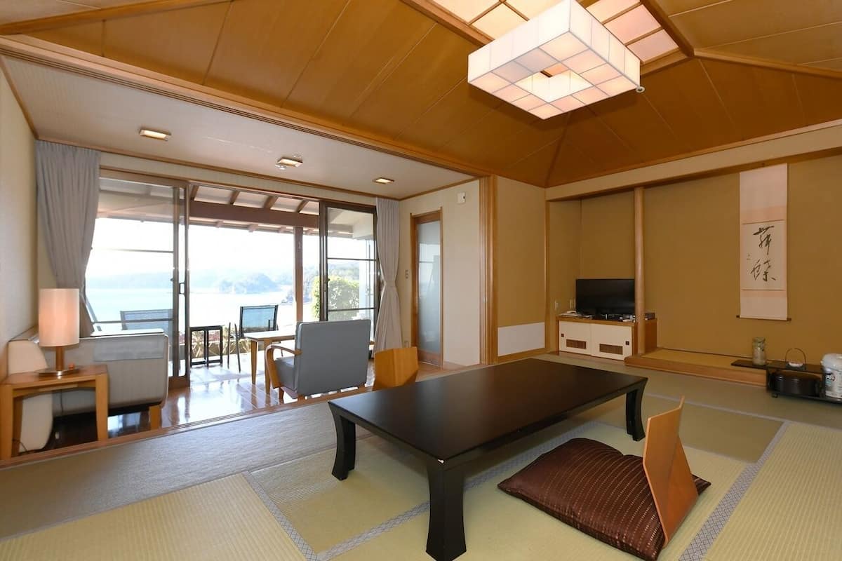 A traditional Japanese-style room with tatami flooring, a low table, a sofa, a television, and a sliding door leading to a balcony with a view.