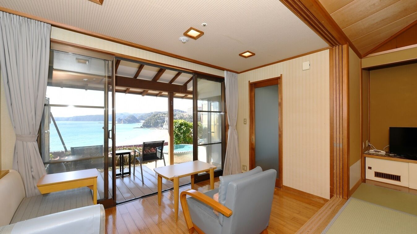 A room with a large sliding glass door leading to a balcony with a view of the sea. There is a small table and a chair inside the room, and a television on a stand.