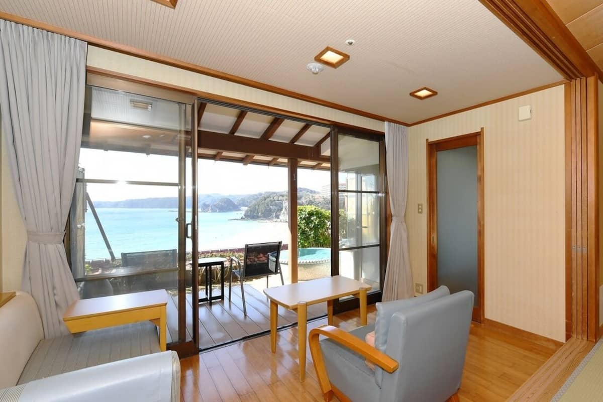 A room with a large sliding glass door leading to a balcony with a view of the sea. There is a small table and a chair inside the room, and a television on a stand.
