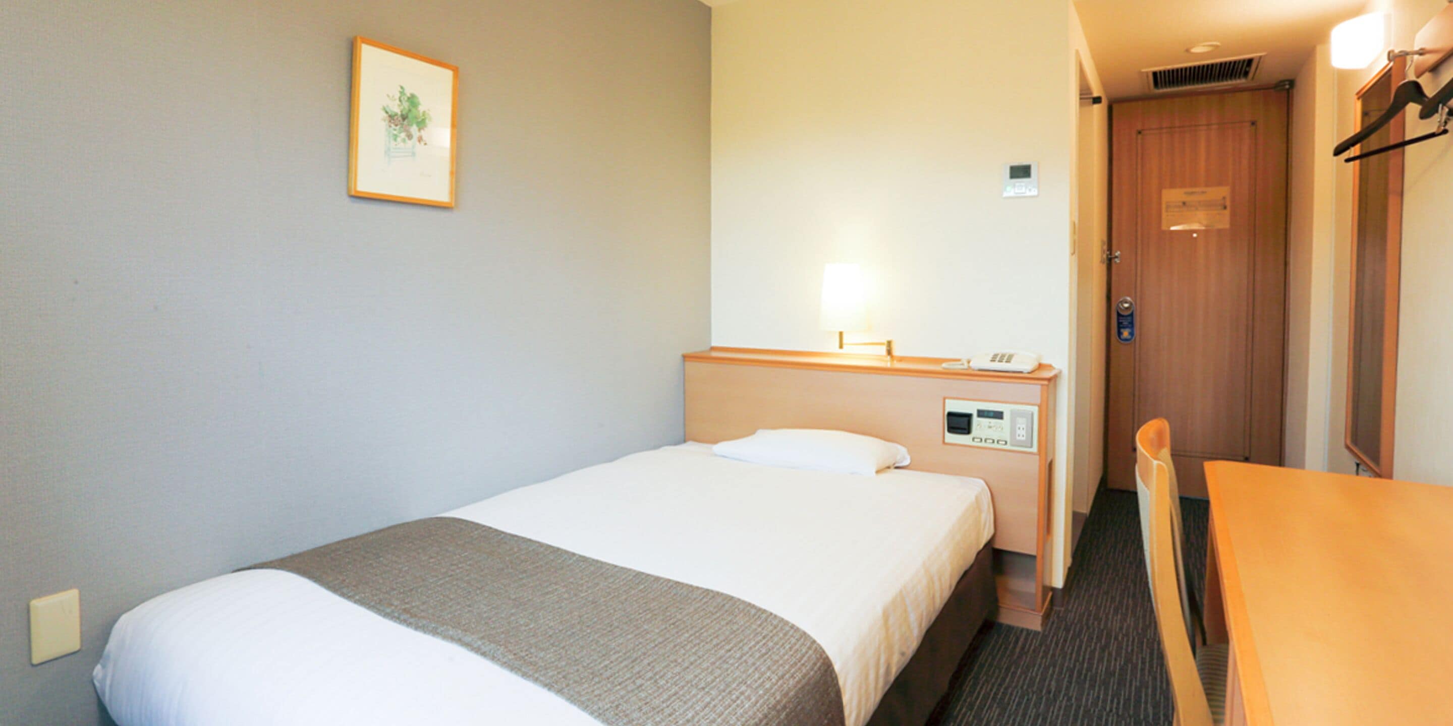 A hotel room with a single bed, a bedside table, a chair, a desk, a lamp, a door, and a framed picture on the wall.