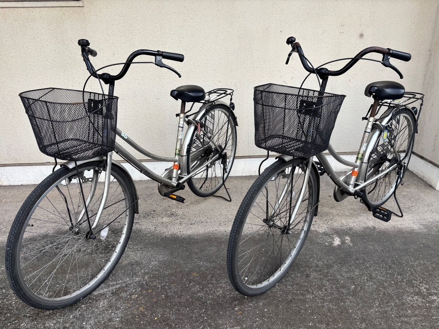 Two bicycles with black wire baskets, parked on a concrete surface against a plain wall.