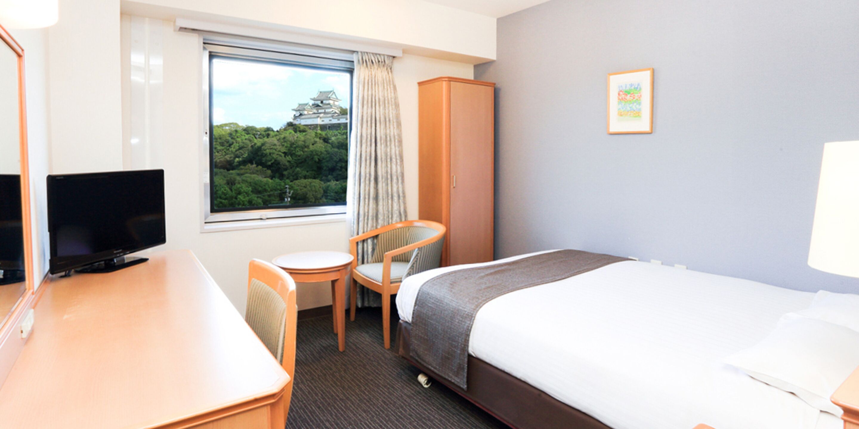 A hotel room with a bed, a desk with a TV, a chair, and a window with a view of trees and a building.