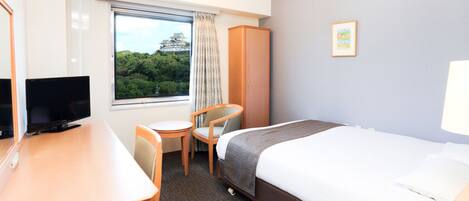 A hotel room with a bed, a desk with a TV, a chair, and a window with a view of trees and a building.