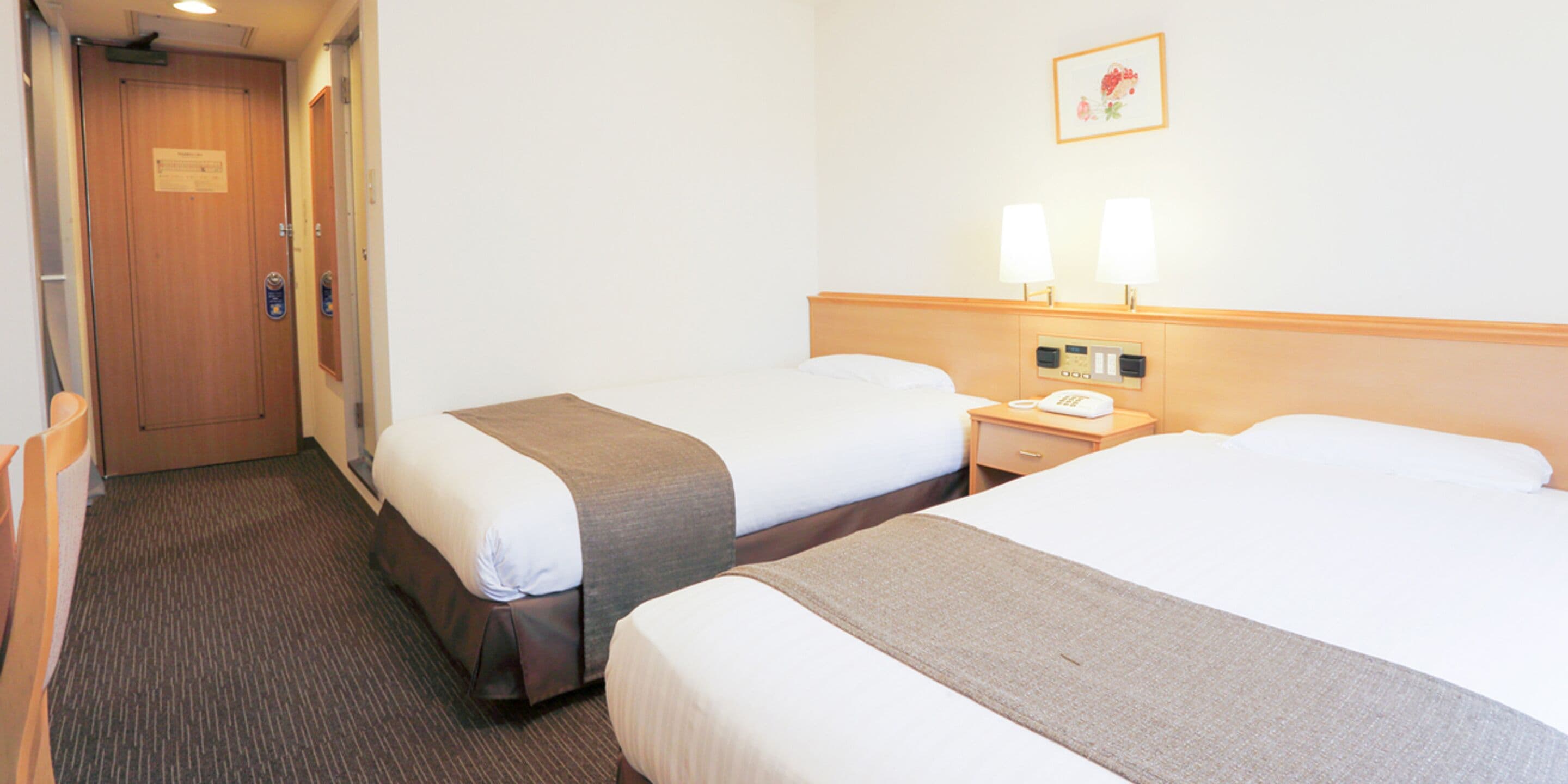 A hotel room with two beds, a wooden door, a bedside table with a phone, and a wall-mounted lamp.