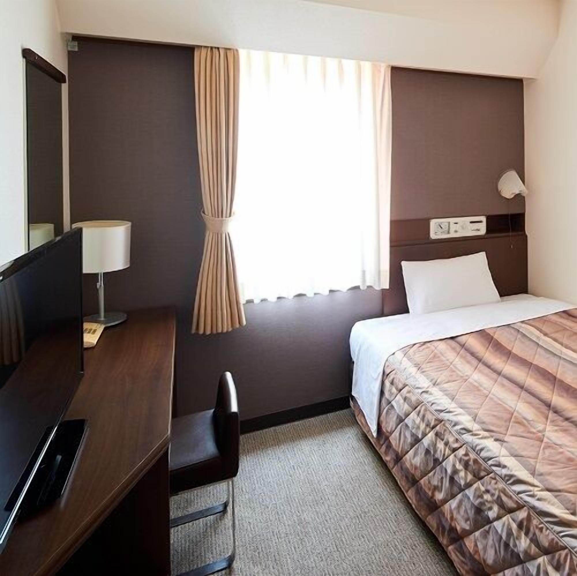 Habitació d'hotel amb llit, escriptori amb làmpada, cadira, televisió i finestra amb cortines.
