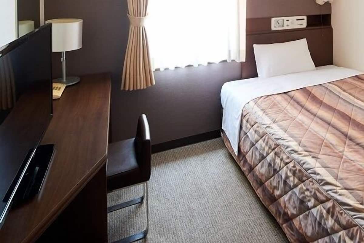 A hotel room with a bed, a desk with a lamp, a chair, a TV, and a window with curtains.