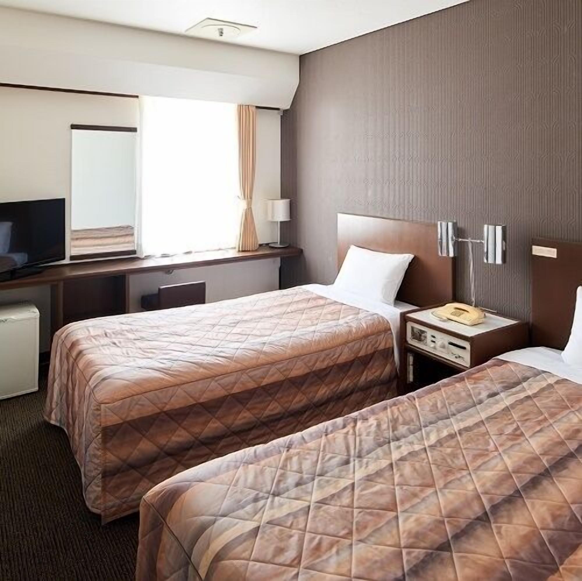 A hotel room with two beds, a desk, a television, and a bedside table with a lamp.