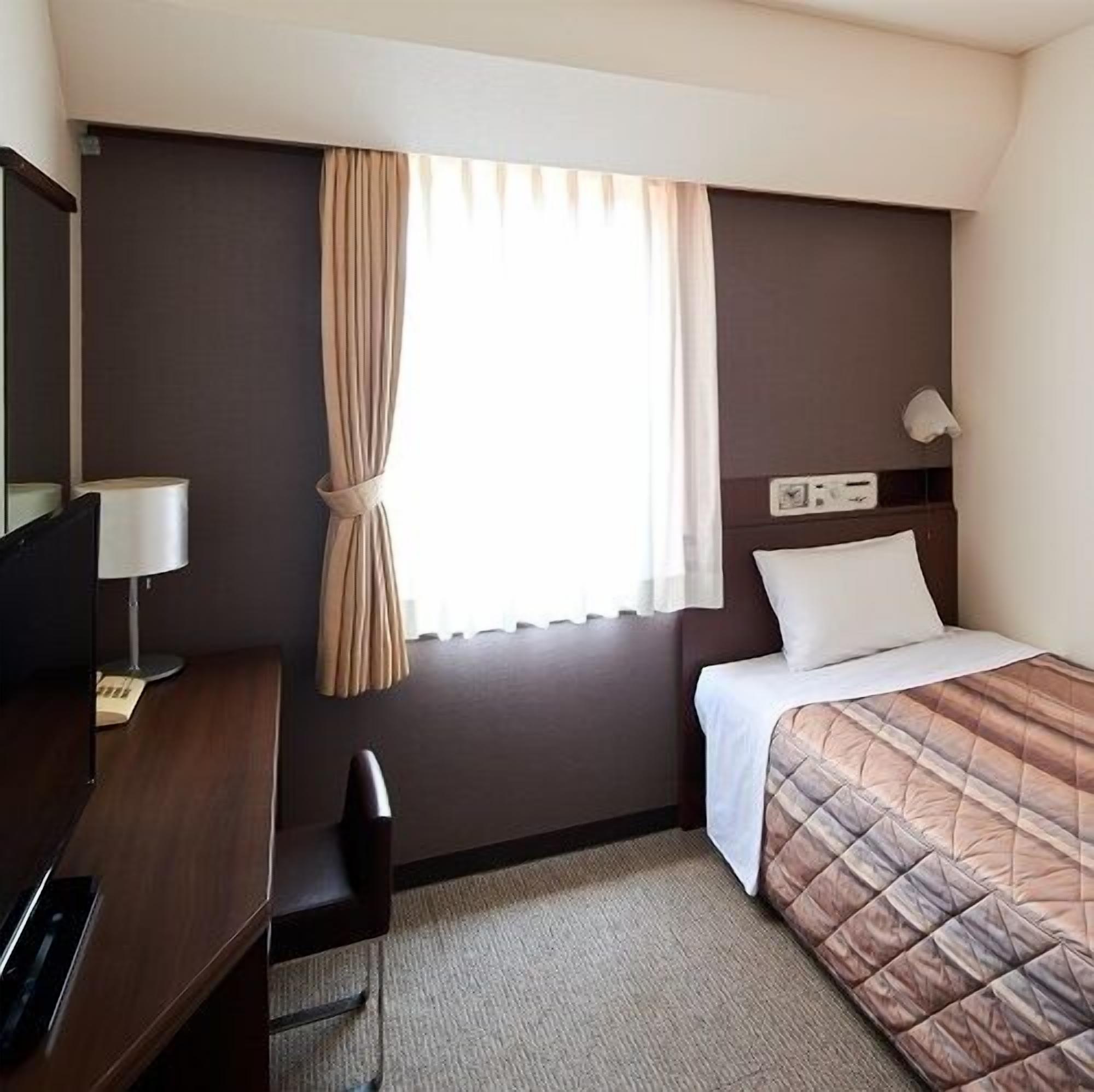 A hotel room with a bed, a desk, a chair, a lamp, and a window with curtains.