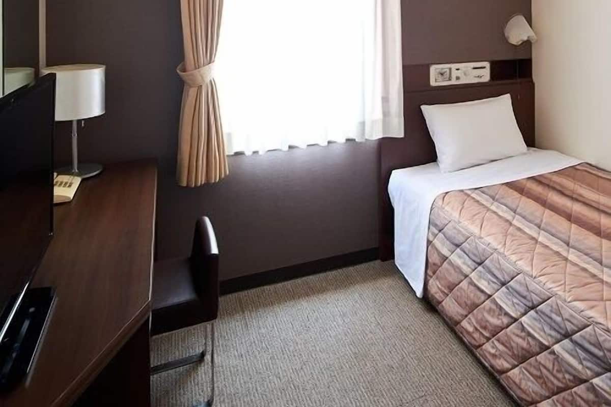 A hotel room with a bed, a desk, a chair, a lamp, and a window with curtains.