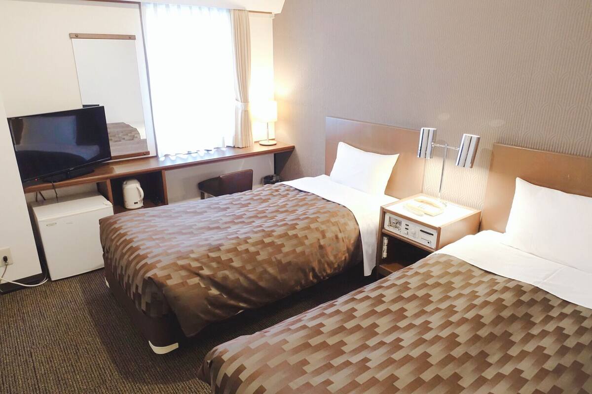 A hotel room with two beds, a desk with a television, a lamp, and a small refrigerator.