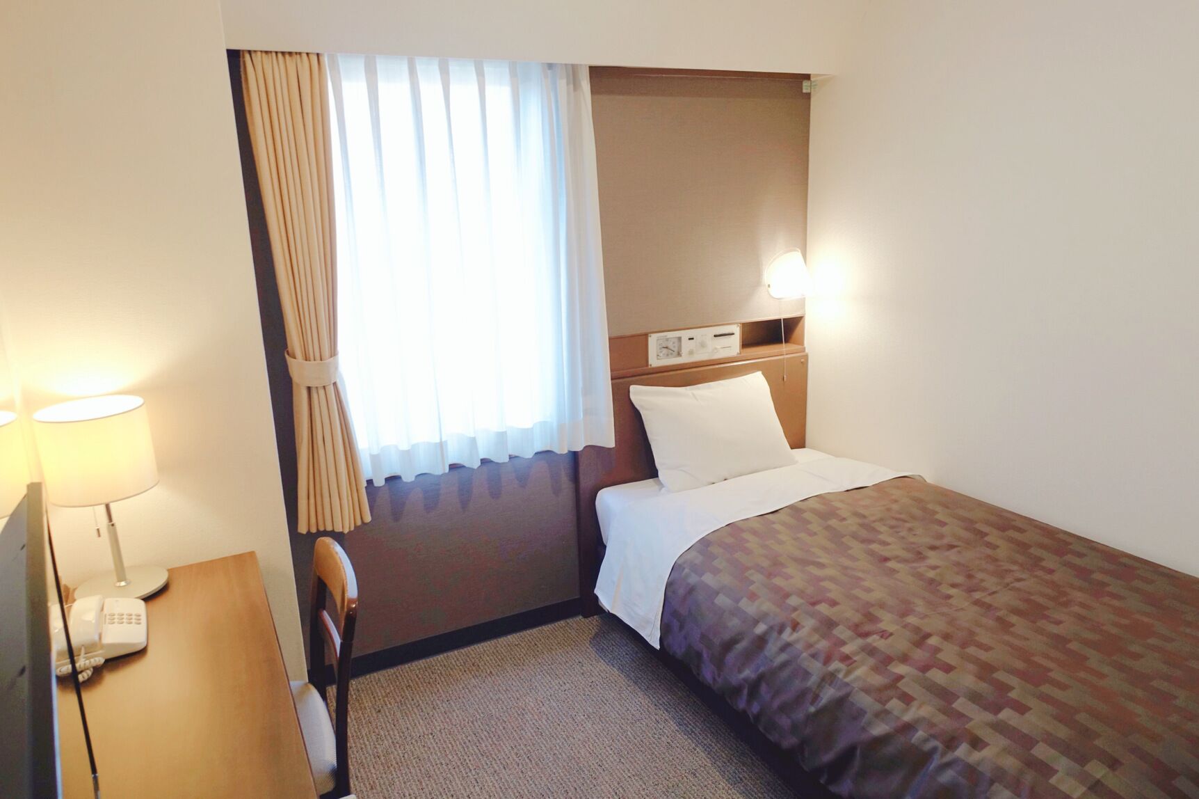 Habitació d'hotel amb llit, escriptori amb làmpada, cadira, finestra amb cortines i panell de control fixat a la paret.