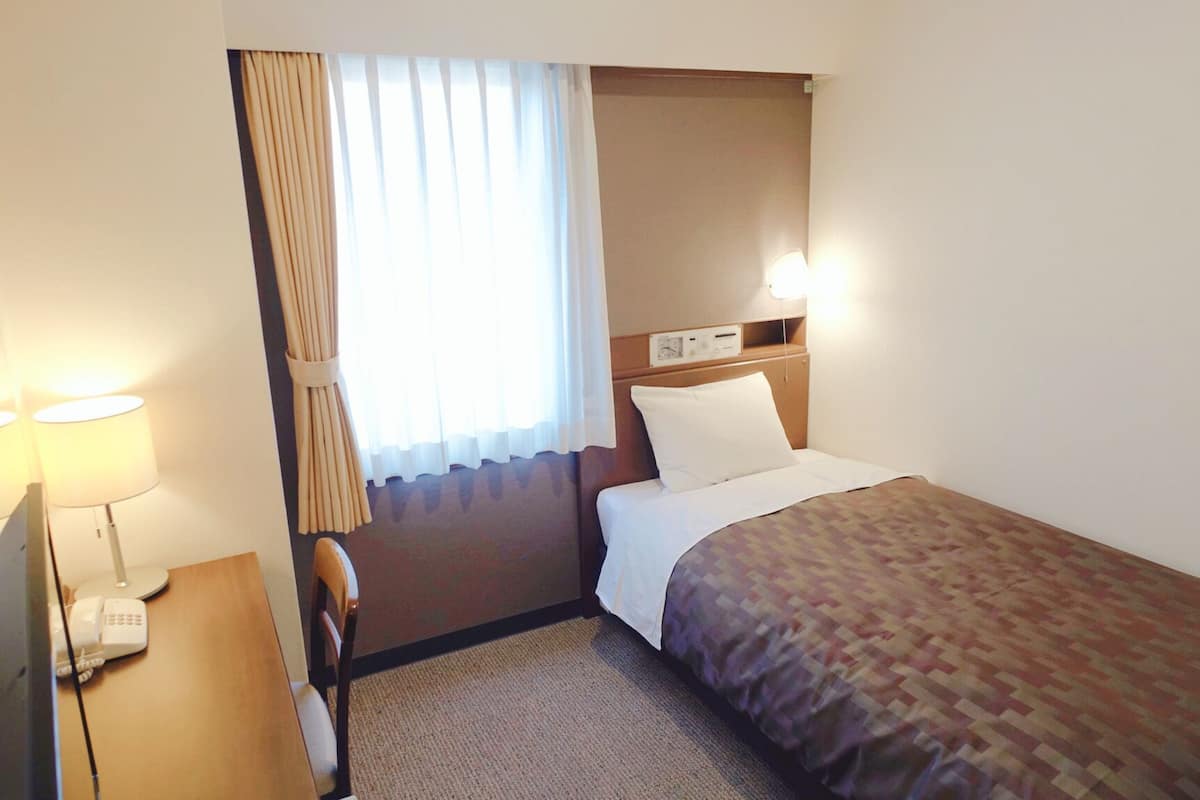 A hotel room with a bed, a desk with a lamp, a chair, a window with curtains, and a wall-mounted control panel.
