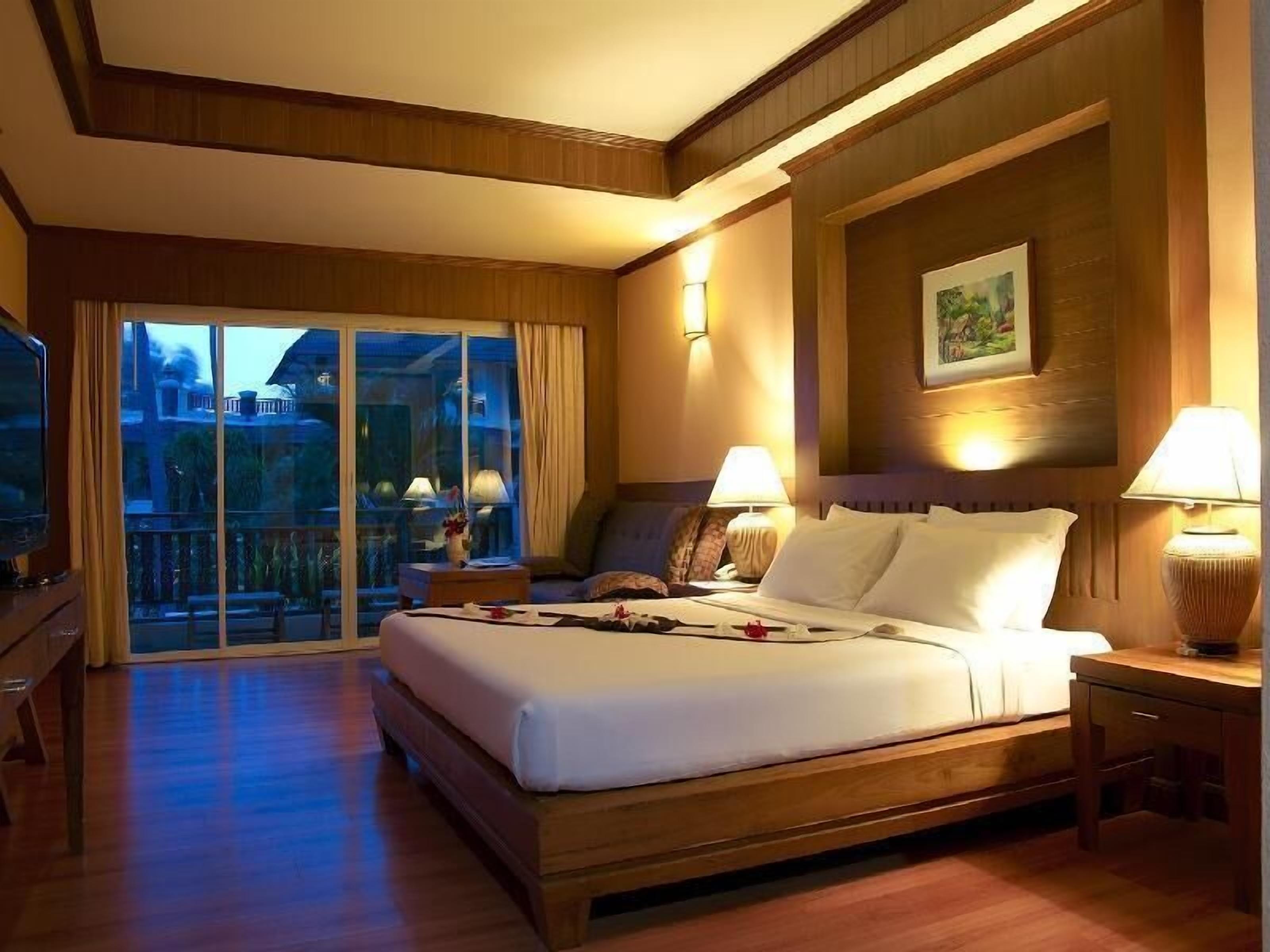 Habitación de hotel con una cama grande, dos mesitas de noche con lámparas, un televisor y un balcón con vistas al exterior.