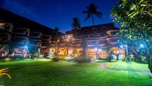A multi-story hotel with a well-lit courtyard, palm trees, and a pathway leading to the entrance. - Aloha Resort (Koh Samui)
