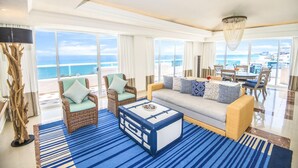 A spacious living room with a large window overlooking the ocean, a blue-striped rug, a wicker sofa, and a dining area with a table and chairs. - Sunscape Cancun Resort & Spa - All Inclusive (Cancun)