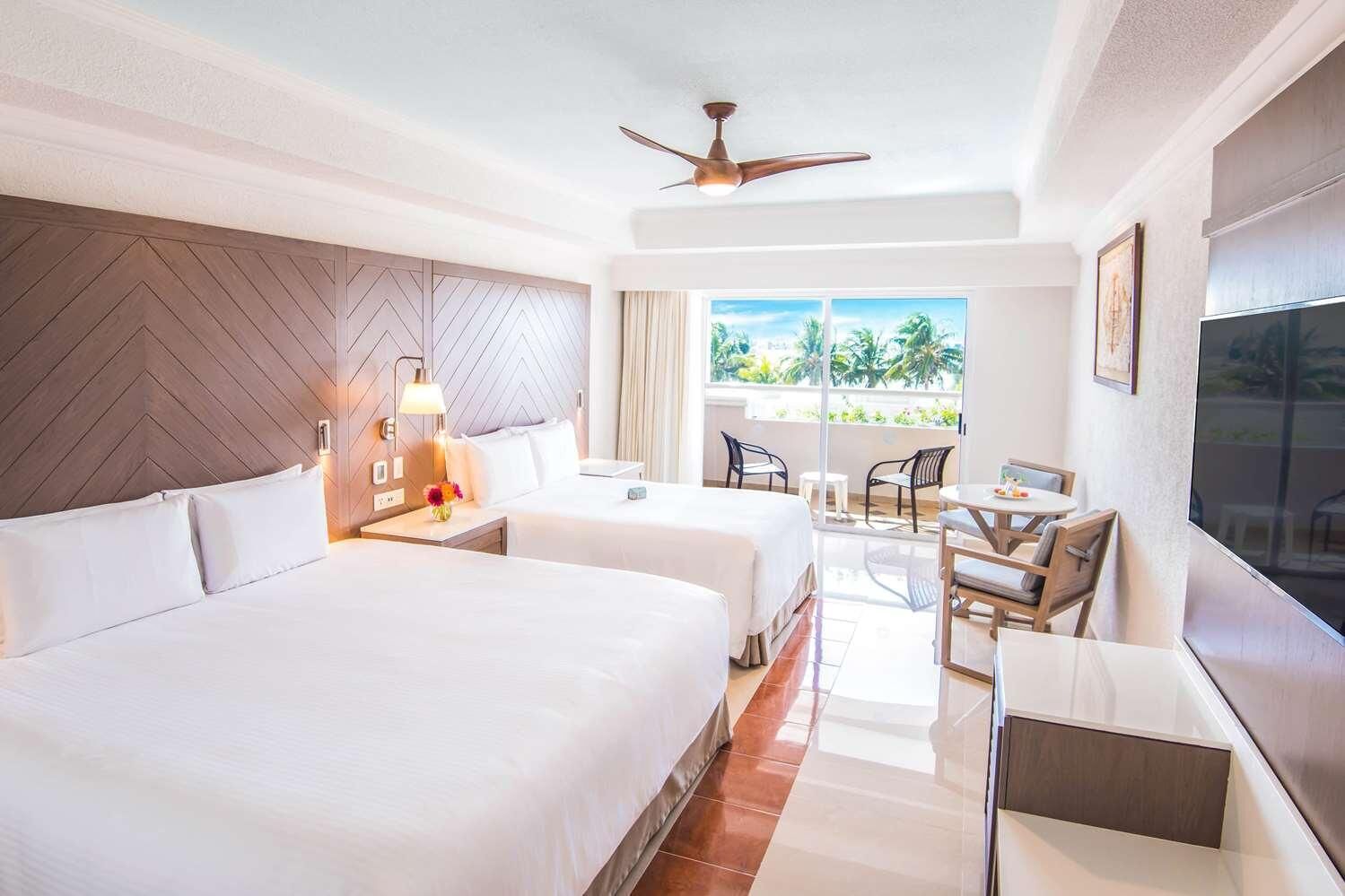 A hotel room with two beds, a desk with chairs, a TV, and a balcony with a view.