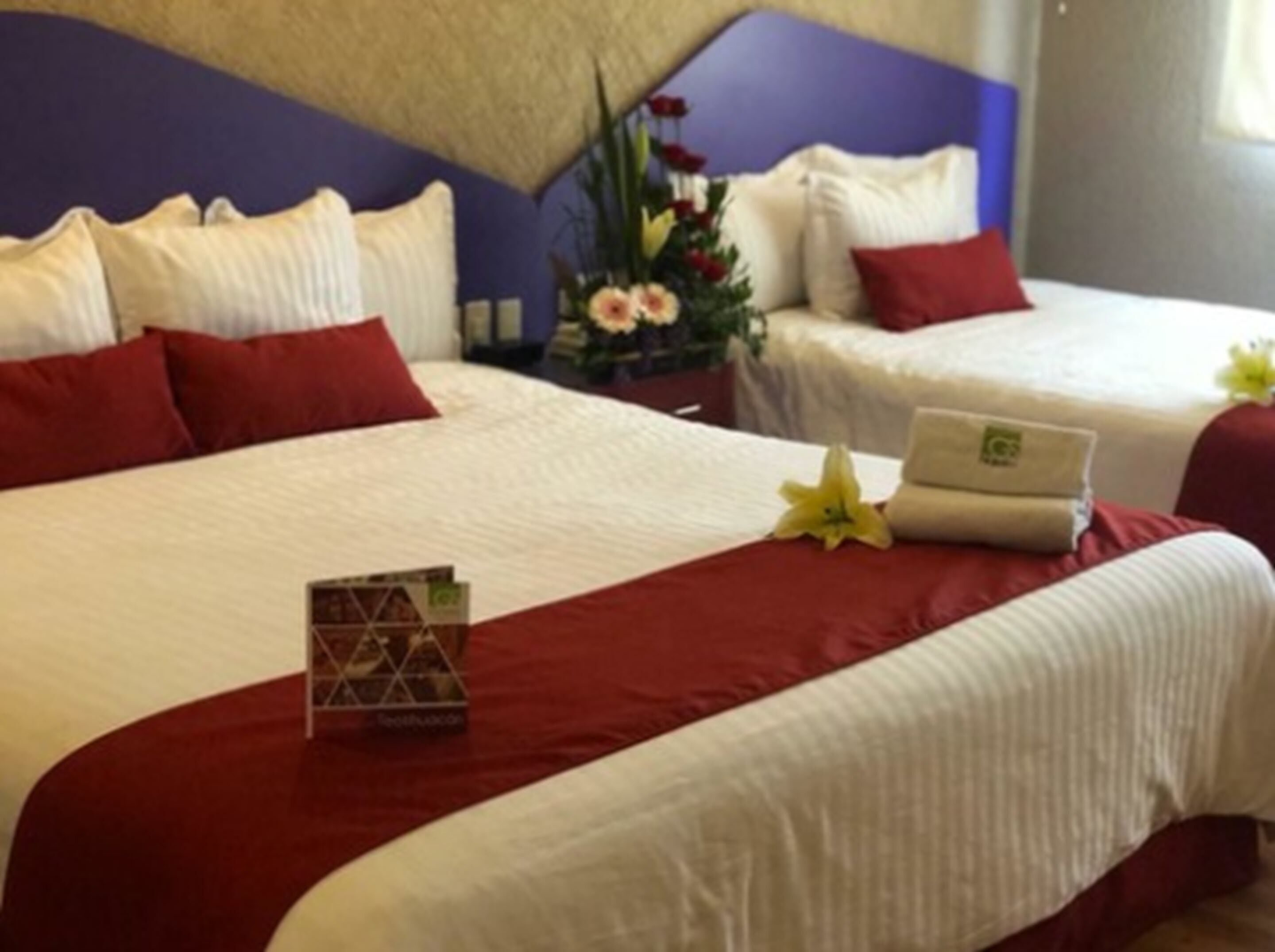 A hotel room with two beds, each with red and white bedding, a nightstand with a lamp, and a floral arrangement on the nightstand.