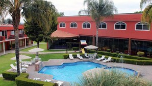 Un hotel con piscina rodeada de sillas de descanso y sombrillas, un edificio rojo con ventanas arqueadas y jardines bien cuidados.