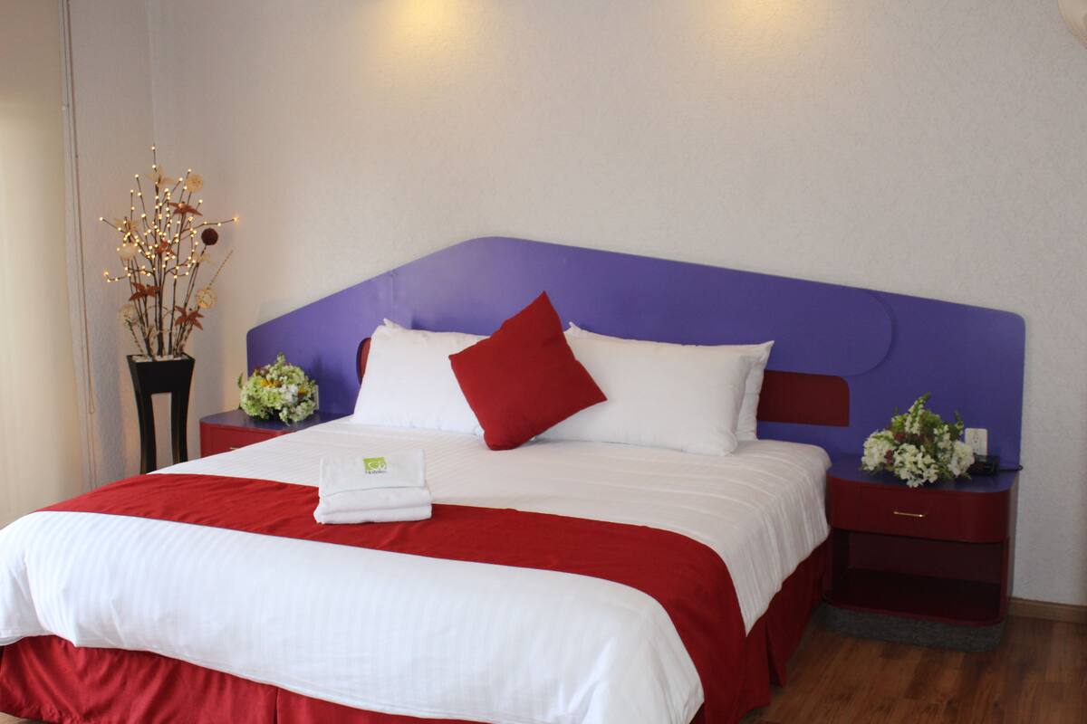 A neatly made bed with a red and white bedspread, a purple headboard, and two nightstands with floral arrangements.