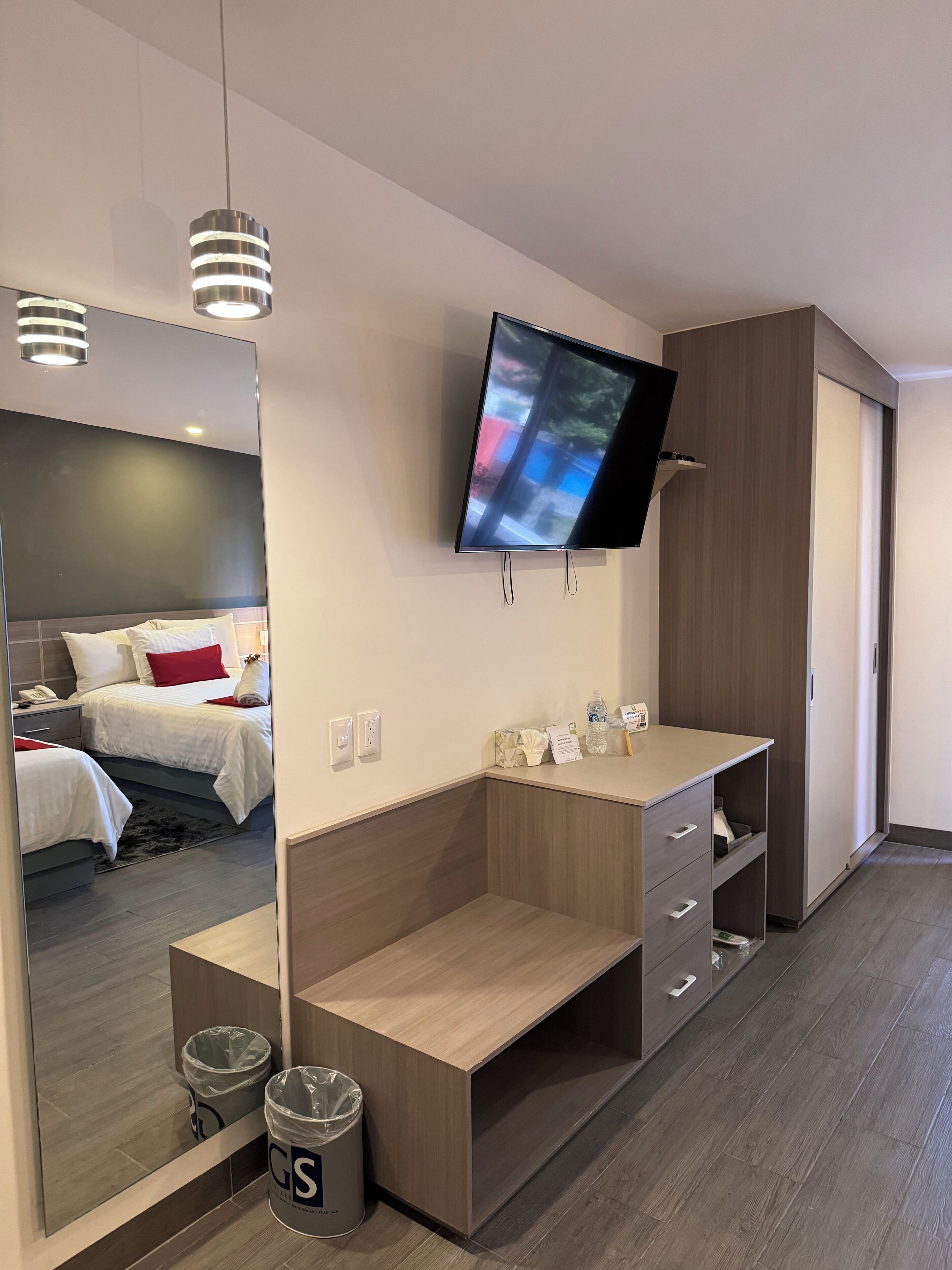 Habitación de hotel moderna con un espejo grande, un televisor montado en la pared, un escritorio de madera y una cama con almohadas rojas.