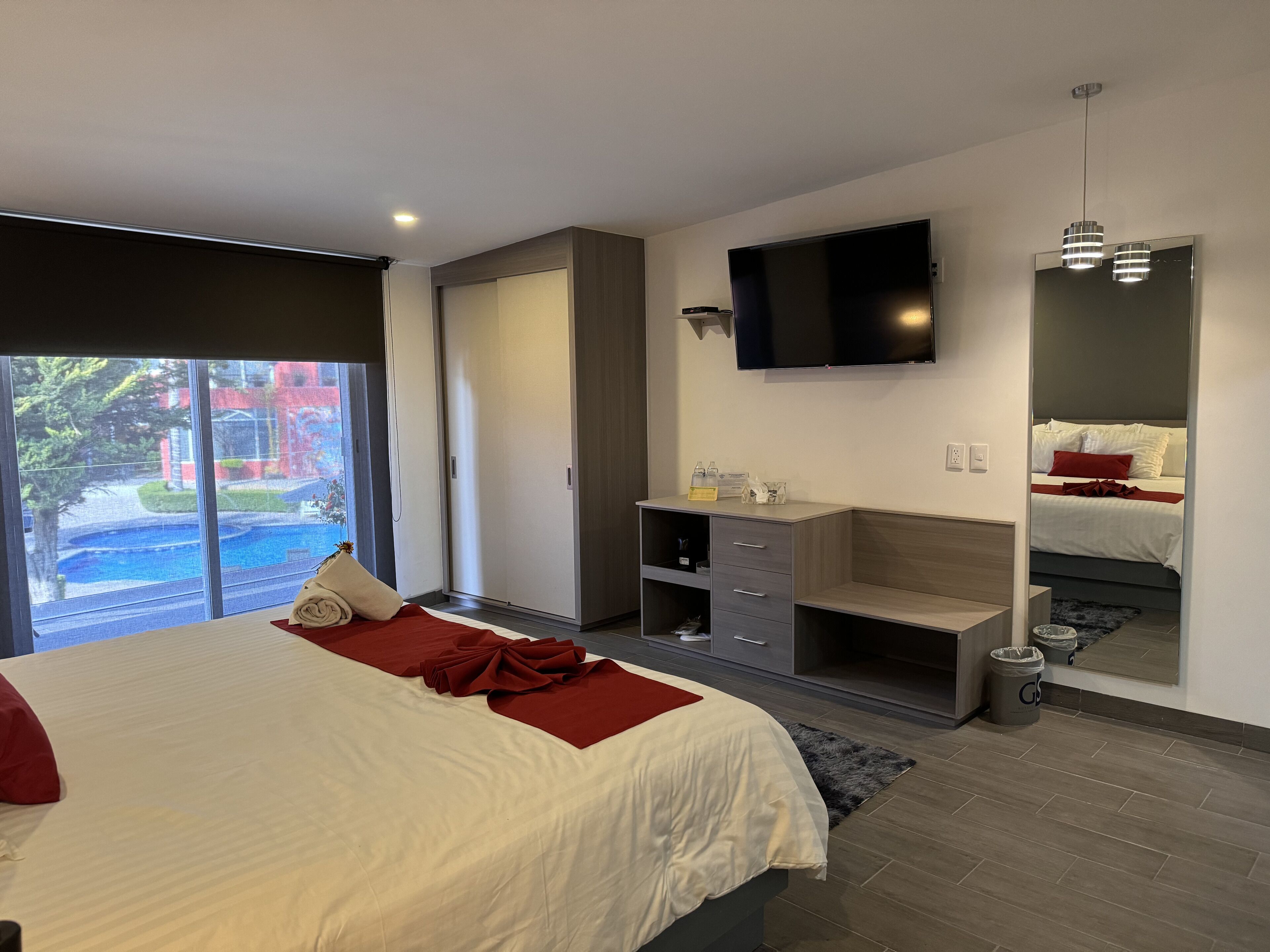 A modern hotel room with a large bed, a TV mounted on the wall, a built-in desk, and a sliding door leading to a pool area.