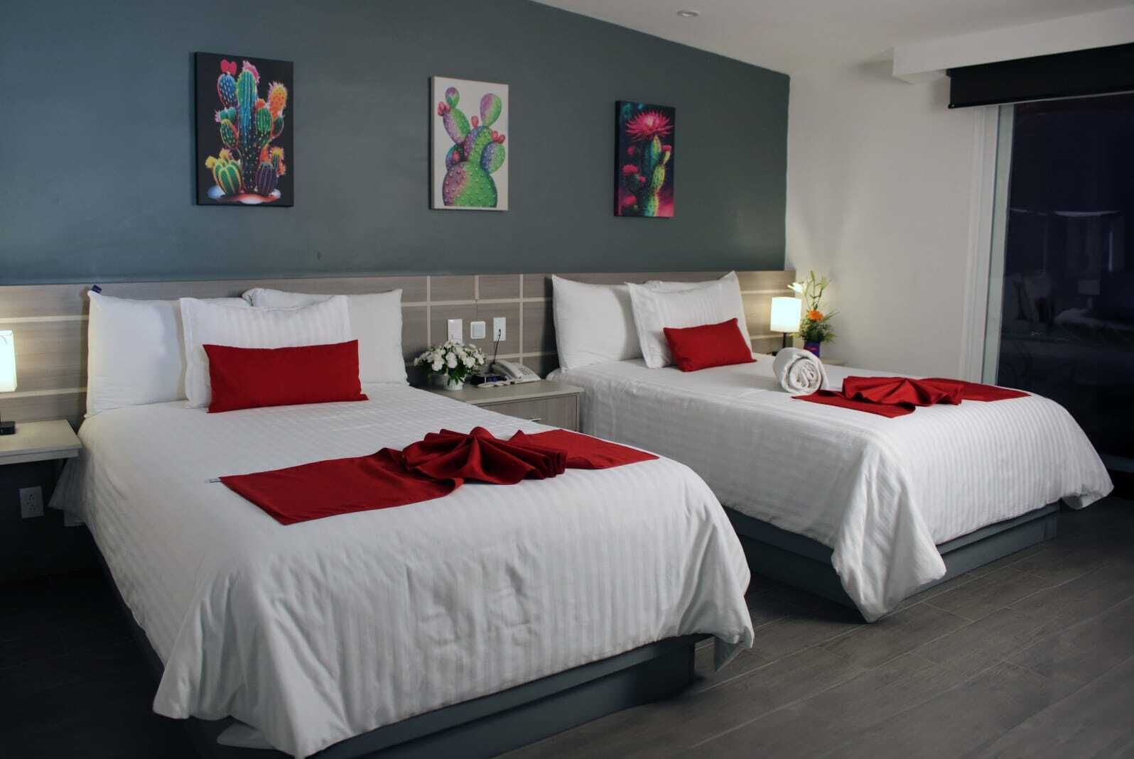 Habitación de hotel con dos camas, cada una con ropa blanca y almohadas rojas. Hay tres cuadros en la pared sobre las camas.