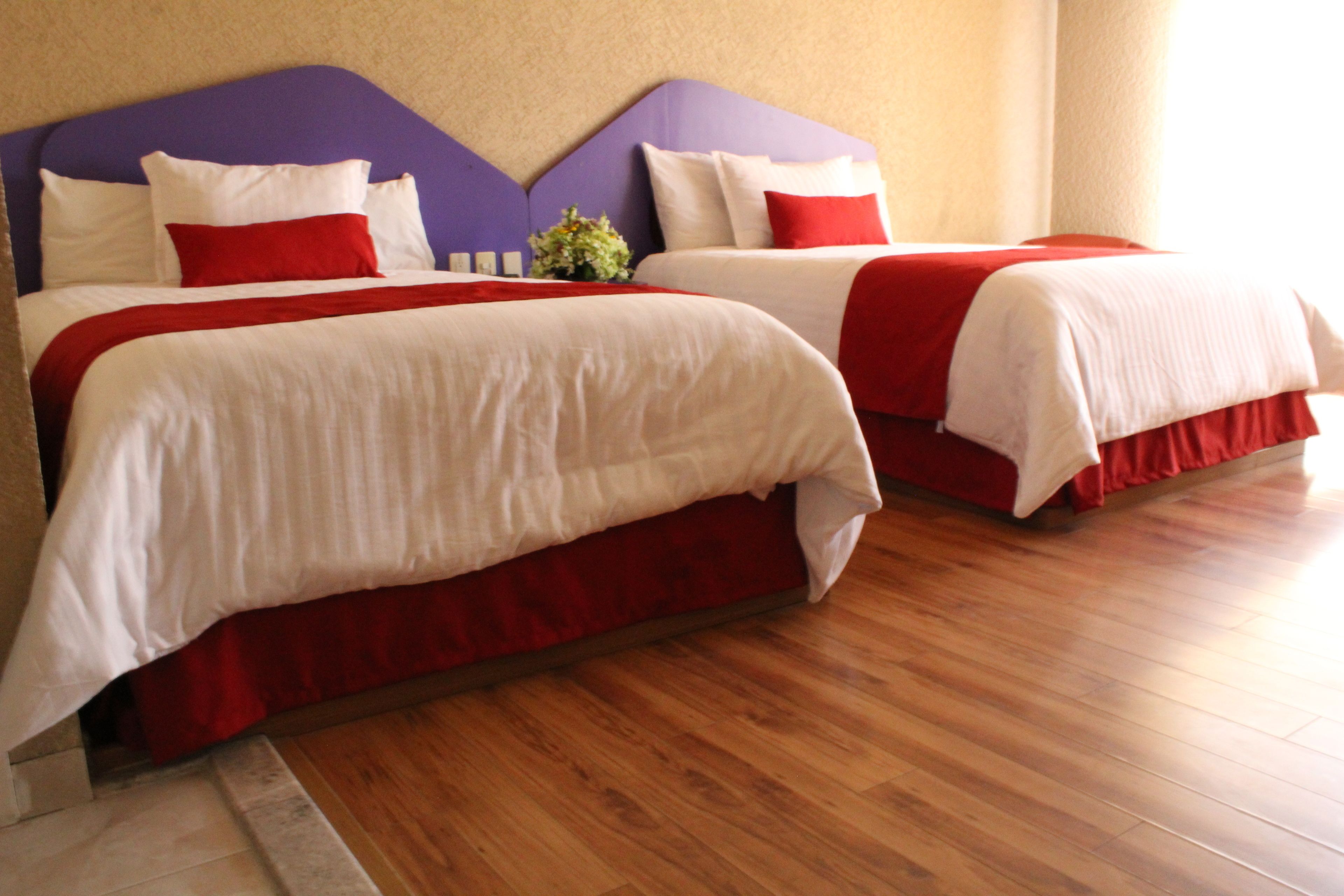 Habitación de hotel con dos camas, cada una con ropa blanca y detalles rojos, cabeceros morados y suelo de madera.
