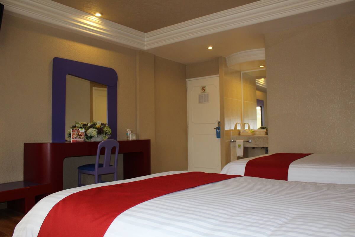 A hotel room with two beds, a desk with a chair, a mirror, and a bathroom visible through an open door.