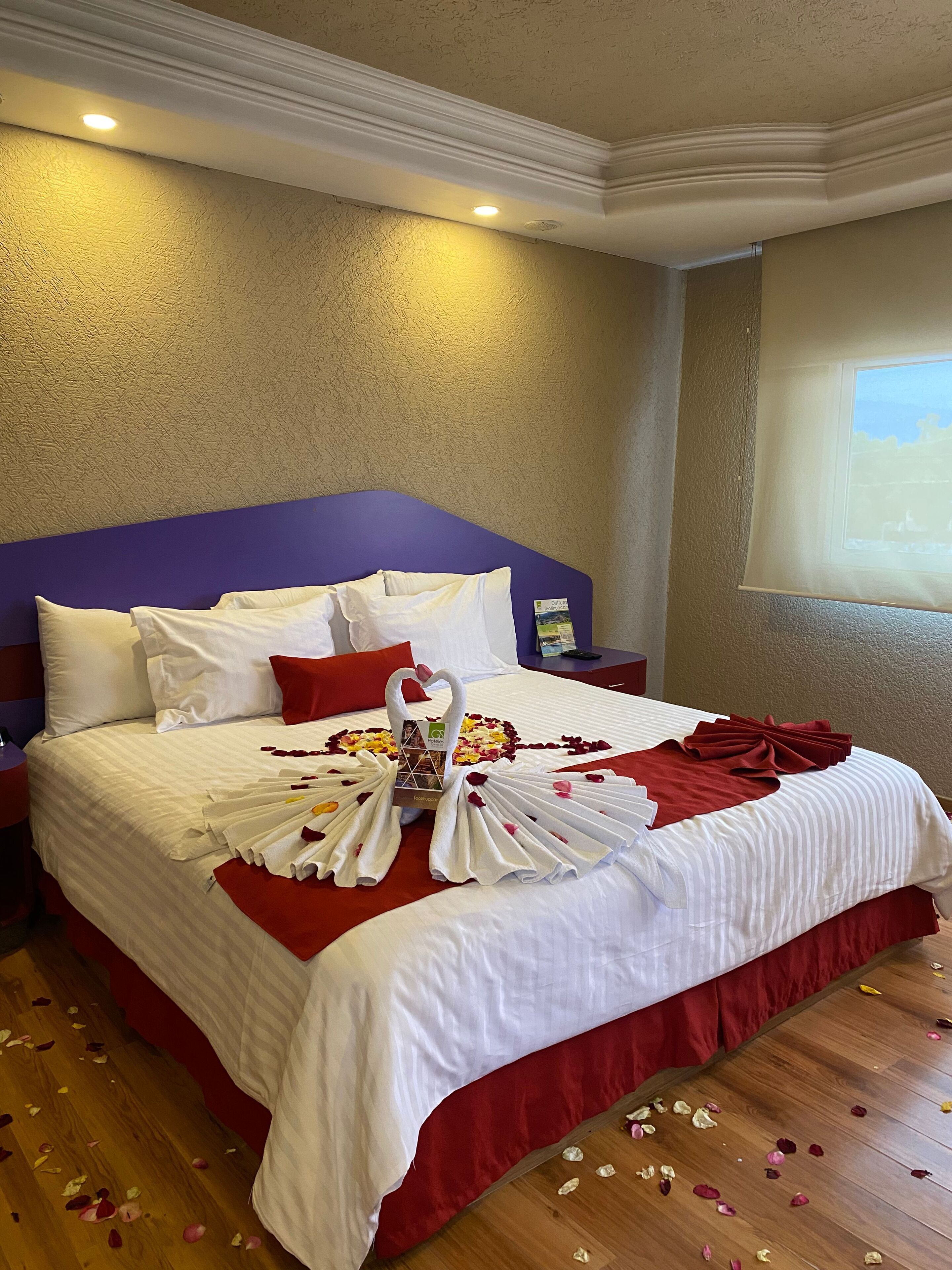 Habitación de hotel con una cama grande, ropa de cama roja y blanca, un cabecero morado y un ventanal con vistas.