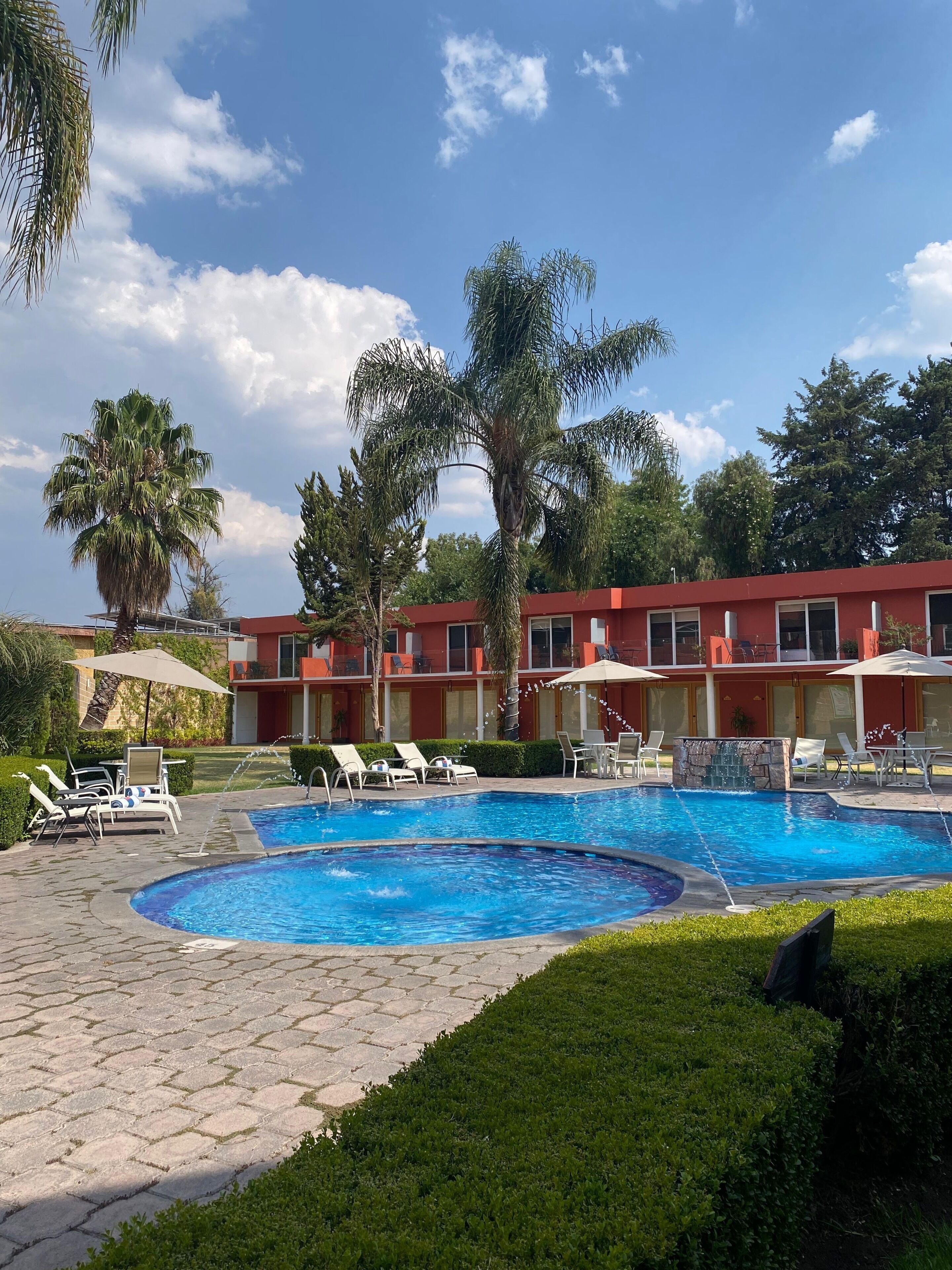 Una piscina con sillas de descanso y sombrillas, rodeada de palmeras y un edificio rojo con múltiples ventanas.