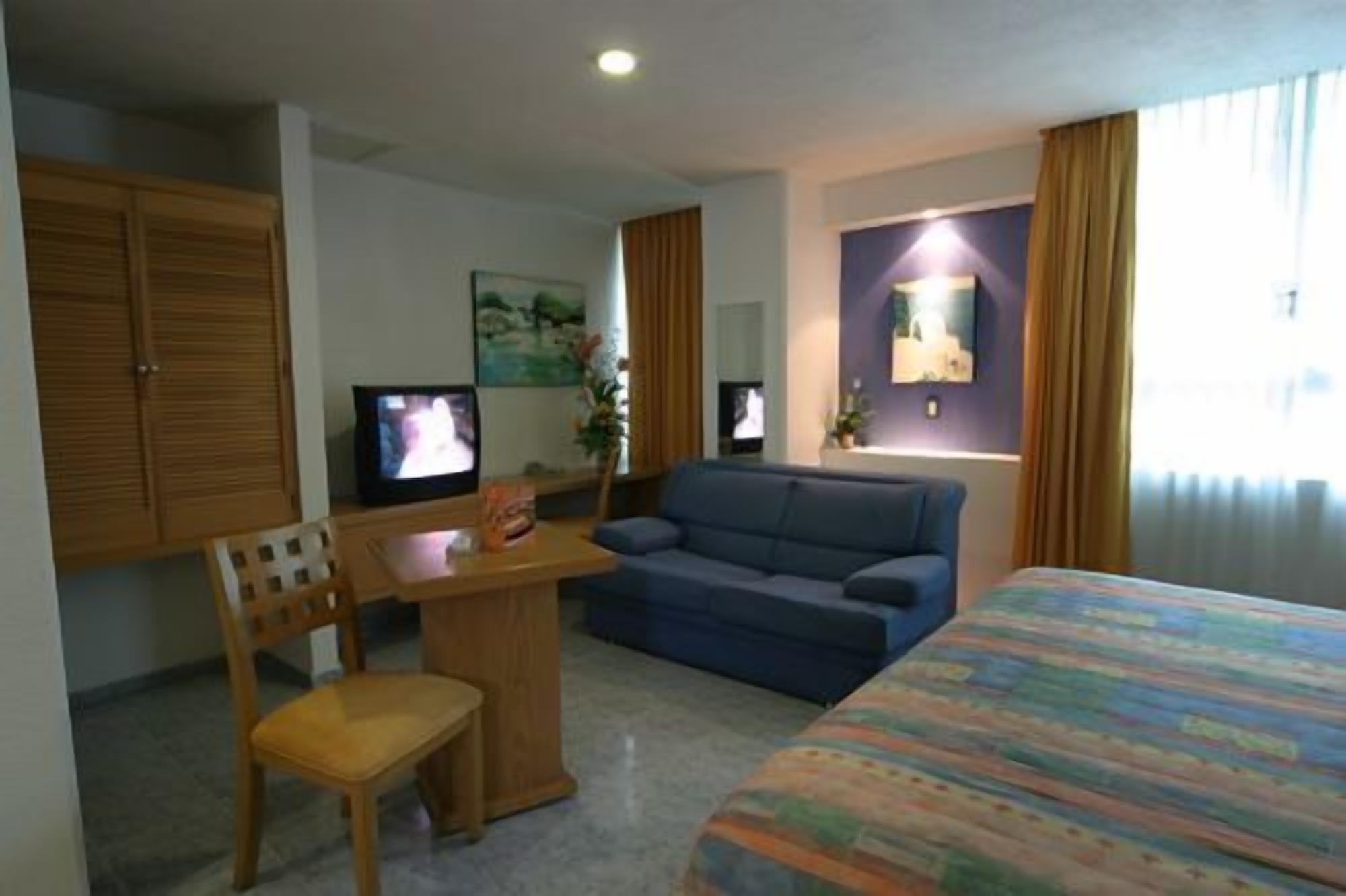 Una habitación de hotel con cama, sofá, escritorio con silla, dos televisores y un cuadro en la pared.