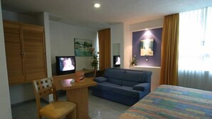 A hotel room with a bed, a sofa, a desk with a chair, two televisions, and a painting on the wall.