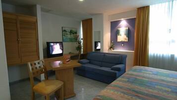Una habitación de hotel con cama, sofá, escritorio con silla, dos televisores y un cuadro en la pared.