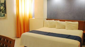 Habitación de hotel con cama, cabecera, mesita de noche, una silla, un cuadro en la pared y ventana con cortinas.