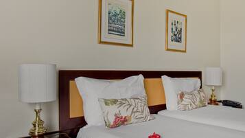 A hotel room with two beds, a wooden headboard, white bedding, a floral pillow, a red decorative item, a nightstand with a lamp, and two framed pictures on the wall.