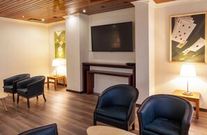Habitación de hotel con televisor de pantalla plana sobre una mesa consola de madera, dos sillones, una mesita de noche con lámpara y un cuadro de cartas de juego en la pared.