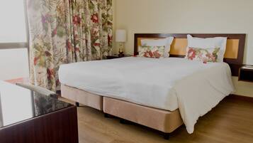Habitación de hotel con una cama grande, cortinas florales, una mesita de noche con lámpara y vistas al exterior.